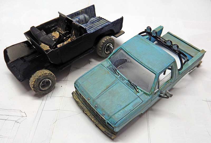 Deux maquettes de pick-up vieillis, l'un noir l'autre bleu patiné, posés sur l'établi
