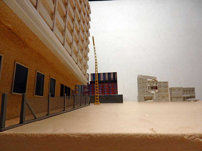 Maquette urbaine avec immeuble, échelle jaune, conteneurs et bâtiments en ruine évoquant The Last of Us