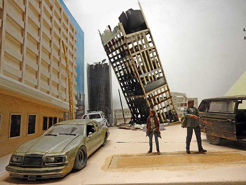 Diorama de ville en ruine avec gratte-ciel effondré, voitures abandonnées et deux figurines de survivants armés