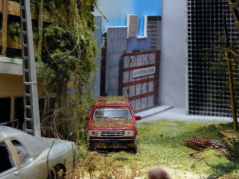 Diorama de rue urbaine post-apocalyptique avec voiture rouge abandonnée envahie par la végétation devant une banque