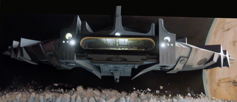 Diorama de l'astronef de Valérian éclairé survolant un sol rocheux devant une planète