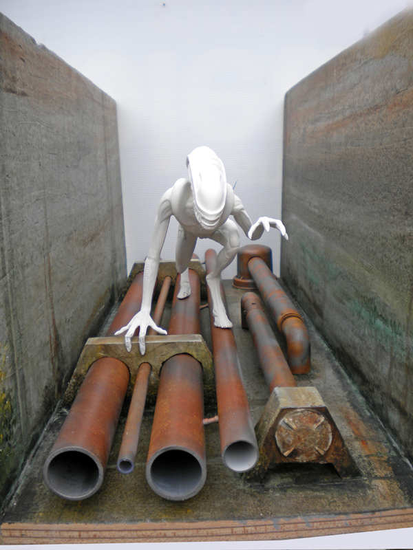 Figurine d'Alien blanche non peinte rampant sur des tuyaux rouillés dans un diorama de couloir