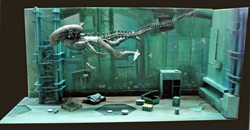 Diorama sous-marin avec un Aqualien squelettique nageant parmi des structures métalliques immergées et éclairées en bleu
