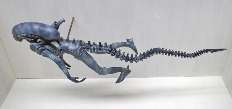 Maquette de créature aqualien bleutée à longue queue segmentée et crâne allongé, vue de profil