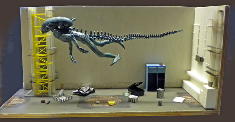 Diorama de maquette montrant une figurine d'alien aquatique à longue queue dans un décor industriel