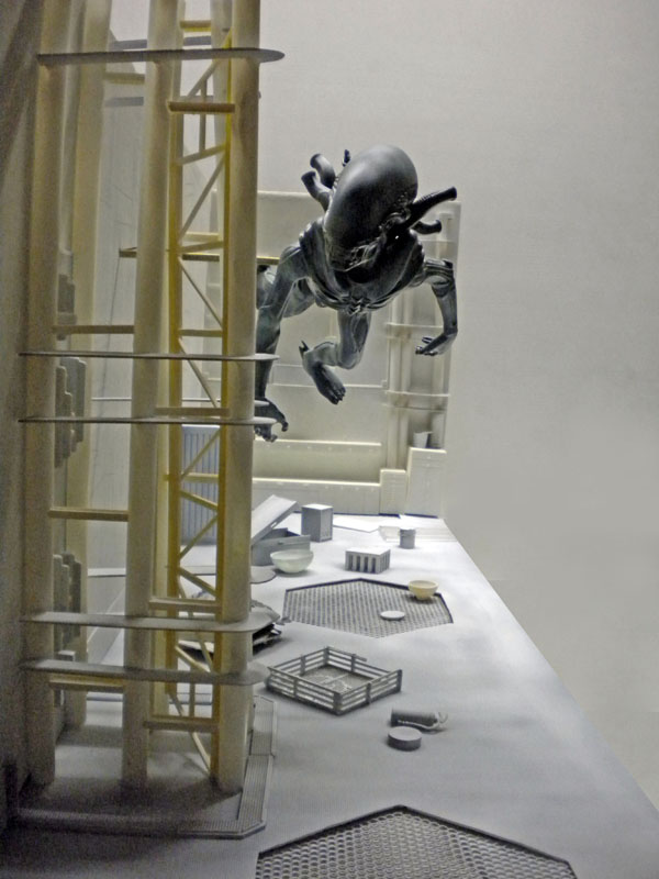 Figurine d'alien noir bondissant au-dessus d'un décor de maquette industrielle gris