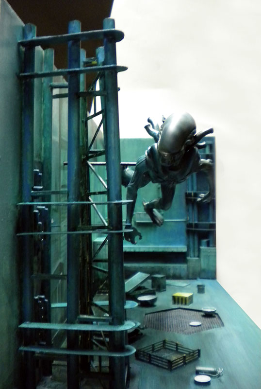 Maquette de créature Aqualien grimpant une échelle métallique dans un décor industriel sombre aux tons bleutés