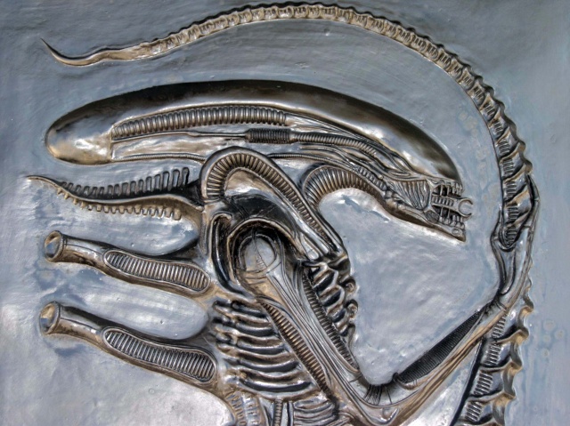 Bas relief patiné gris métallisé montrant la tête allongée de l'Alien et sa longue queue