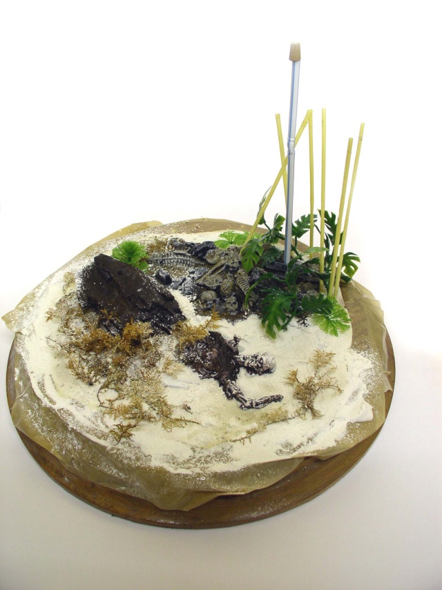 Diorama circulaire de maquette représentant un sol sablonneux avec rochers, végétation et squelette en cours de montage
