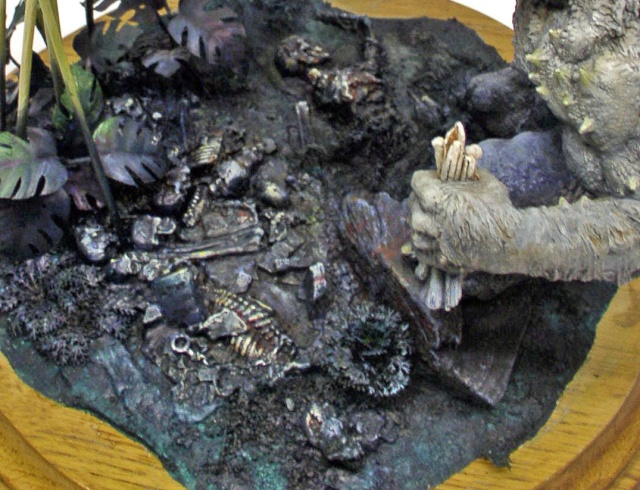 Diorama de maquette montrant un sol jonché de squelettes et la main griffue d'une créature tenant des ossements