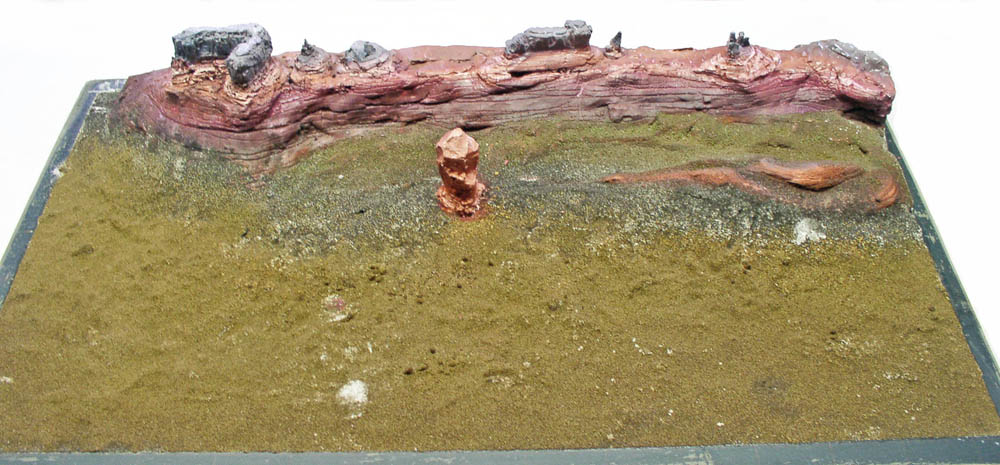 Maquette de diorama martien avec sol sablonneux ocre, falaise rocheuse rougeâtre stratifiée et monolithe cuivré central