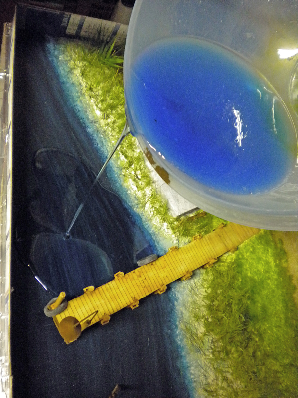 Coulée de résine bleue versée sur un diorama avec ponton en bois et végétation