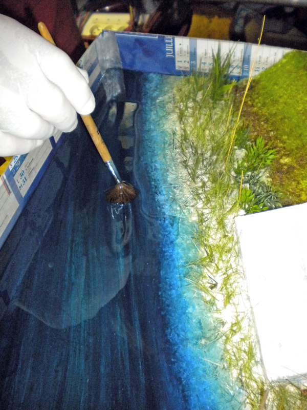 Main gantée appliquant au pinceau la résine bleue de l'eau le long de la berge herbeuse du diorama
