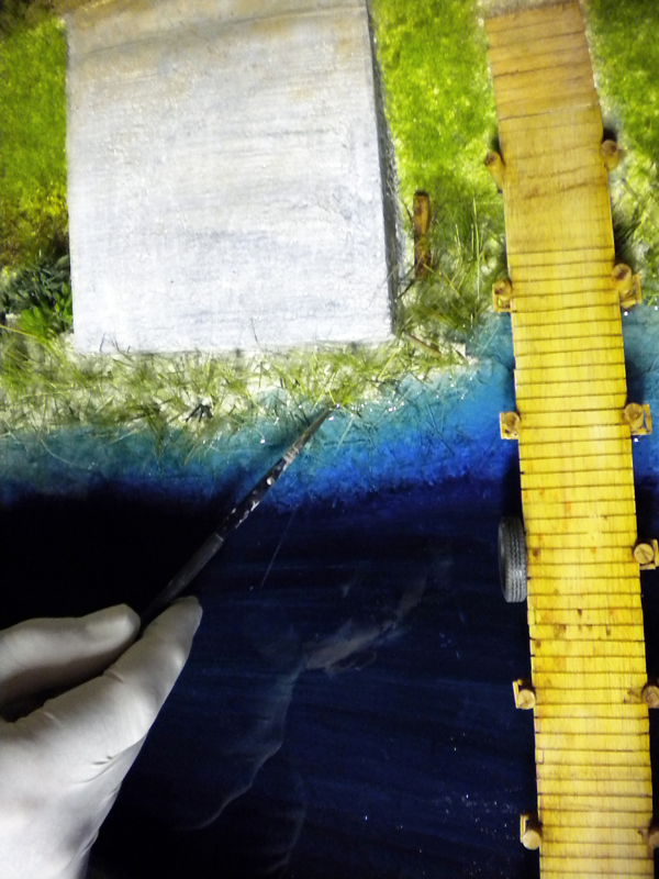 Main gantée appliquant un pinceau sur l'eau en résine bleue d'un diorama bordé d'herbe et d'un ponton