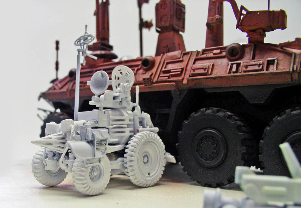 Maquette de petit rover gris à antenne devant un grand transporteur martien rouille à roues