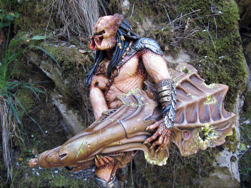 Figurine Predator mâchoires ouvertes tenant une arme organique devant un rocher moussu