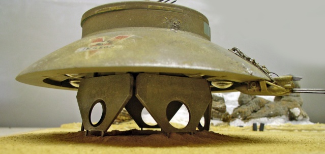 Maquette de soucoupe volante INGSOC posée sur trépied ajouré dans un décor désertique