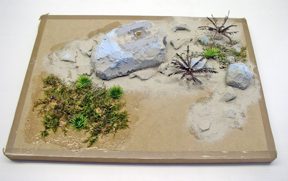 Diorama de maquettisme en construction avec rocher peint, sable, palmiers miniatures et végétation sur socle carton