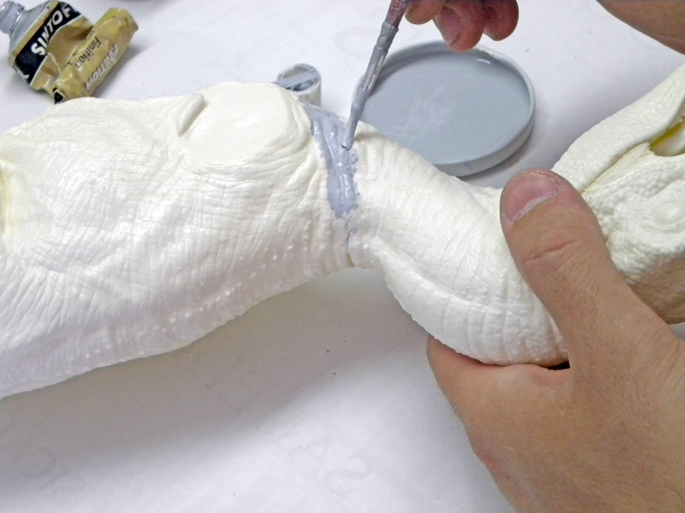 Application de mastic gris au pinceau sur le joint d'assemblage de la maquette Velociraptor tenue à la main