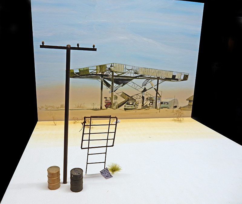 Diorama de maquettisme avec poteau électrique, barils et échelle devant décor désertique de station-service abandonnée